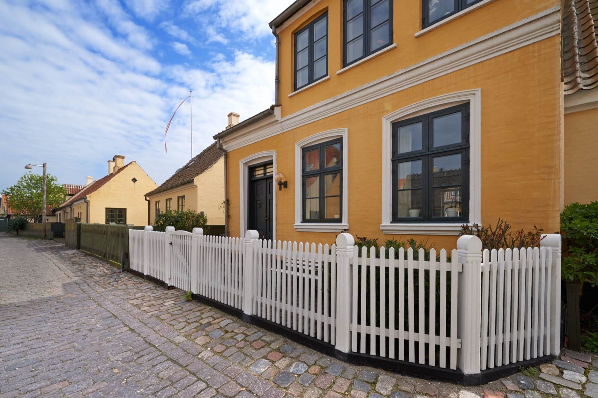 En livfull gul fasad pryder det gamla huset, med dess fönster omramade av svart och omgivet av ett vitt staket, allt placerat på en kullerstensbelagd gata